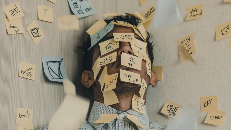 Deprivation & Allostasis - The Cost of Sleep Loss and of Adaptation, The image is a man sleeping against a wall with sticky notes on his head and the walls.